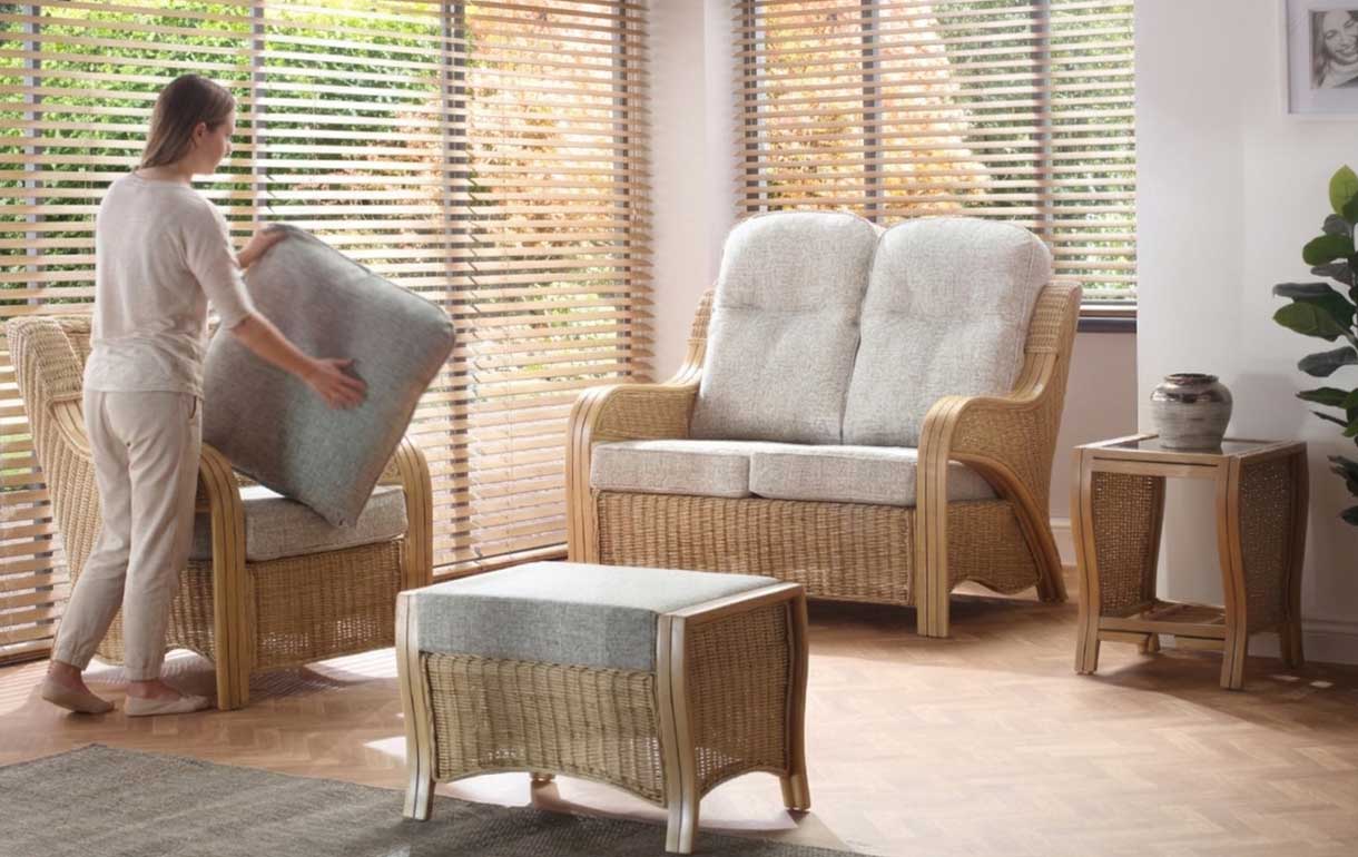 A person maintenance-flipping reversible seat cushions on a cane wicker sofa set in a sunroom with fitted blinds